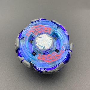 Puede incluir: Un juguete de peonza circular con un anillo exterior de metal plateado y detalles en azul oscuro. El centro muestra una capa azul translúcida con intrincados patrones geométricos rojos que rodean un perno hexagonal plateado central.