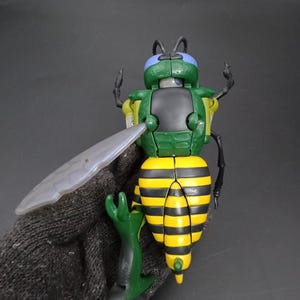May include: A green, yellow, and black toy insect with a clear wing. The toy has a segmented yellow and black abdomen, green head and legs, and black antennae. The toy is held by a gloved hand.