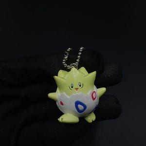 Puede incluir: Figura de Togepi verde claro y blanco con una cadena plateada. El Togepi tiene una cara sonriente, detalles rojos y azules, y mide aproximadamente 5 cm de alto. La figura es un objeto de colección.
