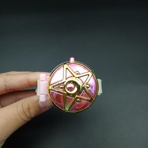以下が含まれることがあります： 星型のデザインのピンクとゴールドの時計。時計には白いバンドとピンクの留め金が付いています。時計は暗い背景に対して手で持たれています。