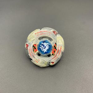 May include: A circular spinning top toy featuring a translucent clear plastic outer ring with red and black flame-like patterns. The centre is a blue hexagonal piece with a white dragon graphic printed on it, surrounded by a metallic silver frame.