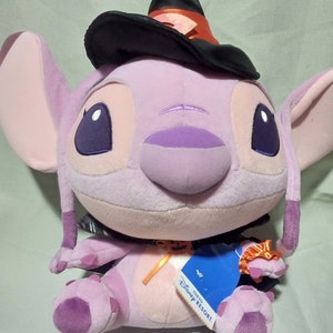 May include: Purple Stitch plush toy wearing a black witch hat with an orange ribbon. The toy is wearing a black and orange costume with a pumpkin patch on the front. The toy has a tag that says "Tokyo Disney Resort".