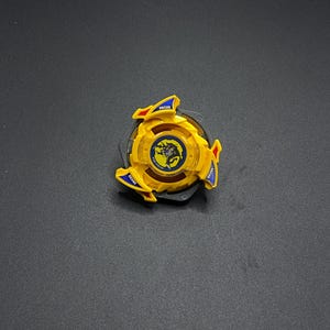 Könnte beinhalten: Ein gelbes und blaues Plastik-Beyblade mit schwarzer Mitte und einem Drachen-Design. Das Beyblade hat den Schriftzug "Draciel" aufgedruckt.