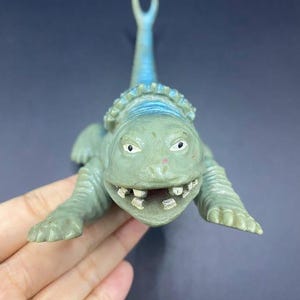 May include: A green and blue toy dinosaur with a wide grin and visible teeth. The dinosaur has a frill around its neck and a curved tail. The toy is made of plastic and has a textured surface.