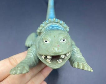 Può includere: Un giocattolo di dinosauro verde e blu con un ampio sorriso e denti visibili. Il dinosauro ha un collare intorno al collo e una coda curva. Il giocattolo è fatto di plastica e ha una superficie strutturata.