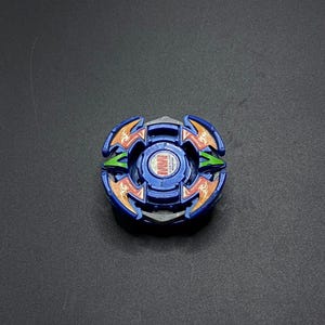 Pode incluir: Um pião azul e prateado com detalhes em laranja e verde. O brinquedo tem um design circular com várias camadas e um emblema central. O pião é projetado para girar e lutar.