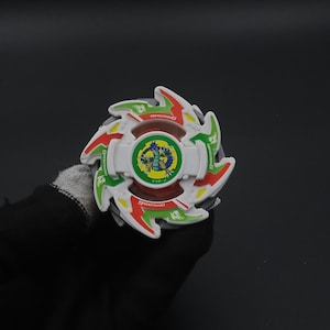 May include: A colourful spinning top toy in white, red, green, and yellow, with the text "DRAGOON G". The centre has a circular design with a blue dragon. The toy is held against a dark background.