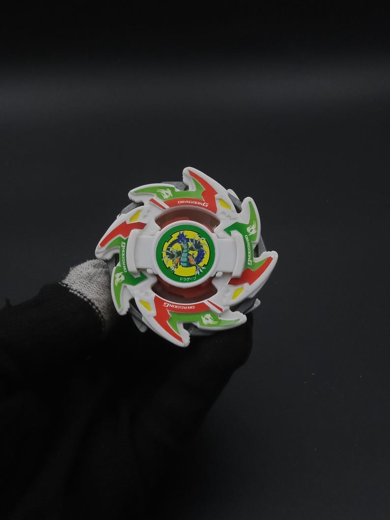 May include: A colourful spinning top toy in white, red, green, and yellow, with the text "DRAGOON G". The centre has a circular design with a blue dragon. The toy is held against a dark background.