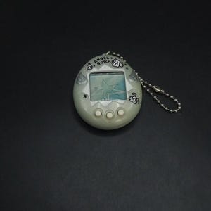 Puede incluir: Un dispositivo de mascota virtual Angel Gotcha! de color blanquecino. El juego portátil presenta una pequeña pantalla que muestra un gráfico de estrella, tres botones de control y una cadena plateada. El texto "ANGEL GOTCHA" está impreso.