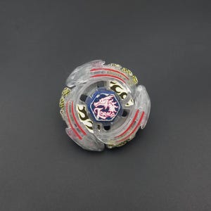 Puede incluir: Un juguete de peonza circular con un anillo exterior de plástico transparente con detalles en rojo y dorado. En el centro, un perno hexagonal azul muestra un gráfico de un dragón blanco y el texto Drago. El diseño incluye tres brazos.