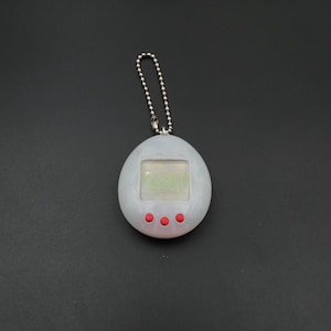 Puede incluir: Un juguete digital para mascotas de color gris claro con una pequeña pantalla y tres botones rojos. El juguete está unido a una cadena de bolas plateada. La pantalla muestra una imagen pixelada. Un juguete retro nostálgico.