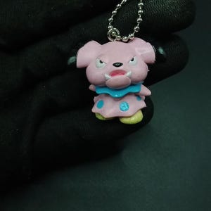 Puede incluir: Un llavero rosa de un personaje de dibujos animados con una cadena de bolas plateada. El personaje tiene una expresión enfadada, con un cuello azul y pies amarillos. El llavero es de plástico.