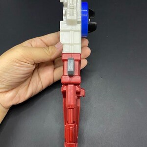 May include: A multi-colored toy gun with a red handle, white body, blue and black details. The toy is held in a hand against a dark background. The toy has a trigger and other mechanical features.