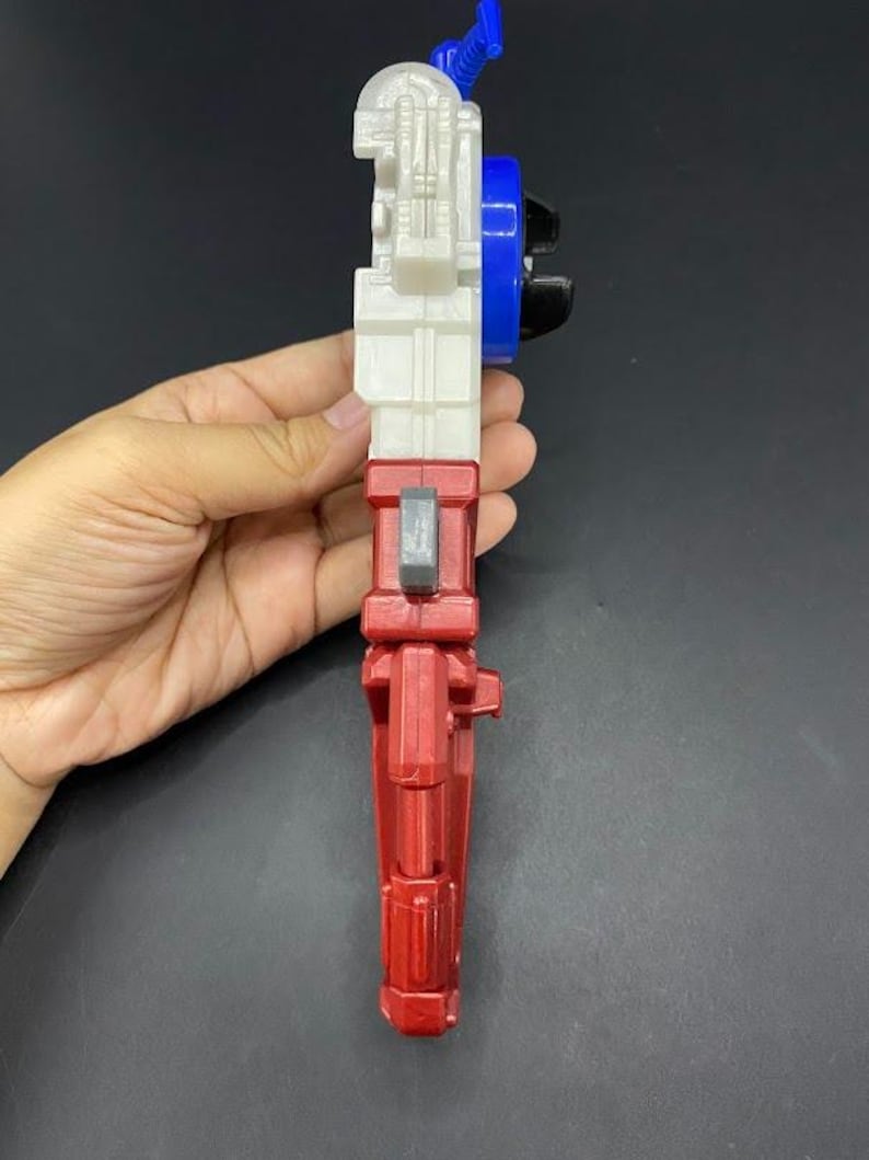 May include: A multi-colored toy gun with a red handle, white body, blue and black details. The toy is held in a hand against a dark background. The toy has a trigger and other mechanical features.