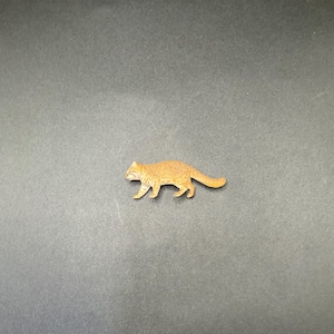 May include: A small, wooden cat figurine in a walking pose. The cat is a light brown color with etched details, and is set against a dark gray background. The figurine is a decorative item.
