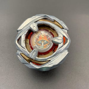 May include: A circular spinning top toy featuring a metallic silver outer ring with gold accents. The centre contains a red translucent component with a graphic illustration of a stylised face and the text BEYBLADE printed in white lettering.