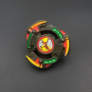 May include: A vintage Beyblade toy, with a circular design and a central emblem. The toy features green, orange, and yellow colours. The word "BEYBLADE" is printed on the green sections. The centre has a red and yellow graphic.