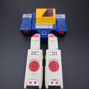 May include: A vintage toy robot with a blue, yellow, and gray color scheme. The robot has a rectangular body with a yellow top and blue sides. Two gray leg pieces with red accents are attached to the body.