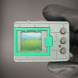 May include: A grey and green digital pet device with a stone-like texture. The screen shows a green field with blue mountains in the background.