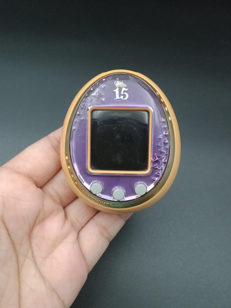 May include: A vintage handheld digital pet device with a purple screen and a gold-colored border. The device has three gray buttons below the screen and the number "15" is displayed on the screen. The device is held in a hand.