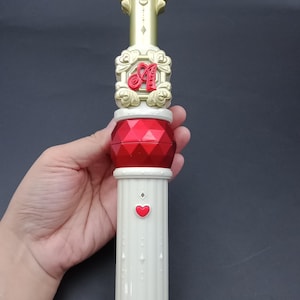 Puede incluir: Una varita decorativa con mango y detalles dorados, un centro rojo en forma de gema y una base blanca en forma de columna. La varita presenta una letra "A" roja y un detalle en forma de corazón. La base tiene un borde dorado.