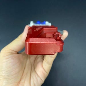 May include: A red plastic toy with white and blue accents. The toy is rectangular with a series of raised sections and a small, protruding element on one side. The object is held in a hand.