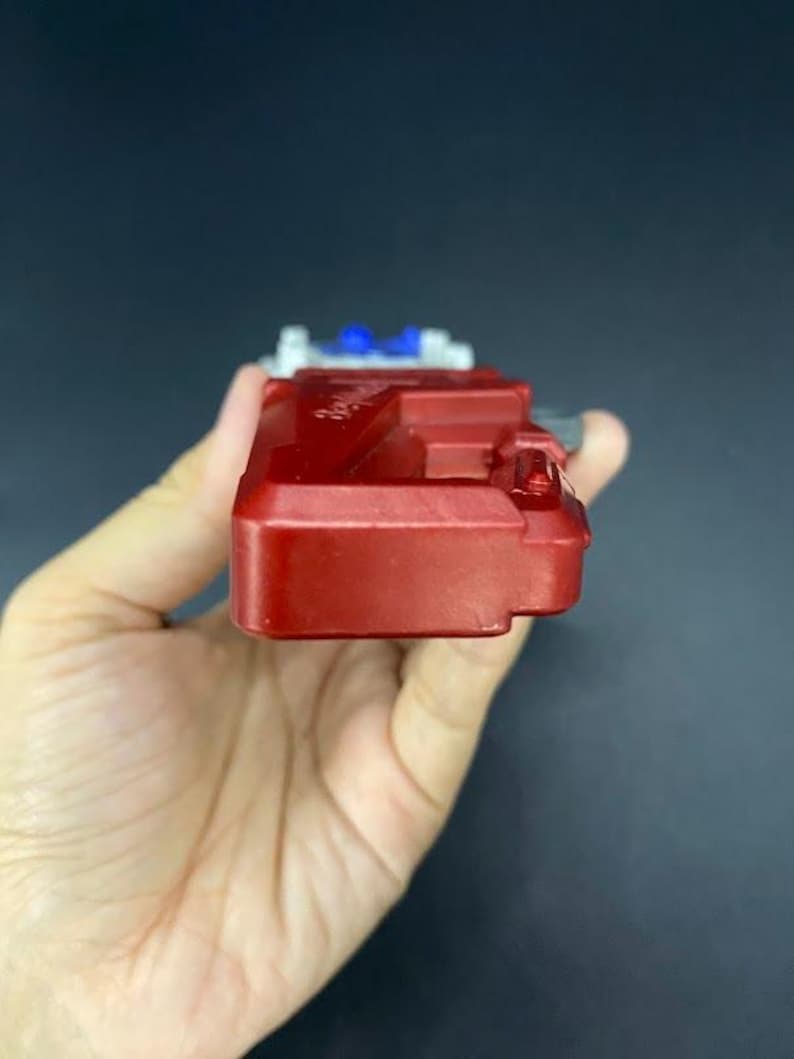 May include: A red plastic toy with white and blue accents. The toy is rectangular with a series of raised sections and a small, protruding element on one side. The object is held in a hand.
