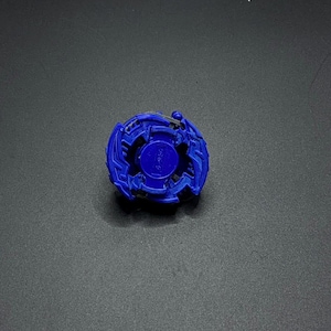 May include: A blue plastic Beyblade with a black center and a silver ring around the center. The Beyblade has a dragon-like design.