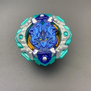 Può includere: Una trottola circolare con un emblema centrale blu raffigurante una creatura a tre teste, circondata da un anello esterno color argento e verde acqua. Il giocattolo presenta accenti gialli visibili sotto lo strato blu e borchie argentate.