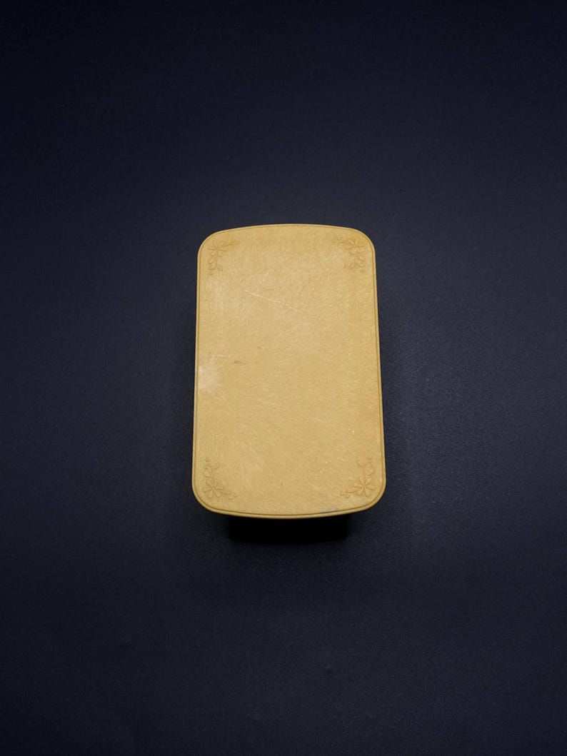 May include: A rectangular, light yellow box with rounded corners. The box has a slightly textured surface and delicate floral designs in each corner. The background is a solid, dark blue.