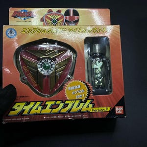 May include: A red and gold plastic toy shield with a green gem in the center. The shield has the words "HAL PANGEP" printed on it. The toy is part of a set that includes a small, clear plastic capsule with a gold and silver toy inside. The packaging is red and gold and features the words "Time Emblem Version-F" and the Bandai logo.