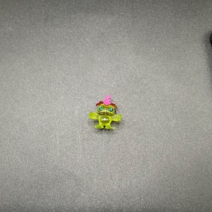 May include: A small, translucent green figurine of a creature with large eyes and a pink hat. The figurine has small arms and legs and is set against a grey background.