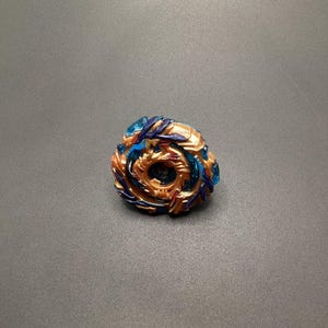 May include: A gold, blue, and purple spinning top toy with a dragon design. The top has a clear blue plastic piece on the outside edge.