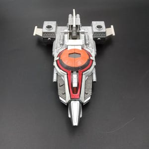 Può includere: Un'astronave giocattolo argentata e rossa con un cerchio arancione centrale e due ali laterali grigie. Il giocattolo ha un design futuristico con pannelli dettagliati e una parte anteriore appuntita, su sfondo nero.