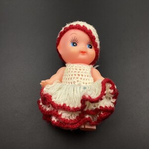 May include: A small plastic doll wearing a white and red crocheted dress and hat. The doll has blue eyes and a pink face.