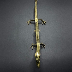 May include: A gold-colored metal lizard figurine with a detailed, textured body and a pointed tail. The lizard is posed with its legs outstretched and its head tilted upwards.