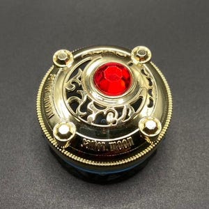 May include: Gold-coloured compact with a red jewel in the centre, surrounded by a black and gold filigree design. The compact has the words "Sailor Moon" inscribed on the bottom.
