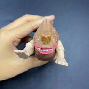 May include: A brown, smiling monster toy with a textured head and light brown arms and feet. The monster has a wide, red-lipped smile with white teeth. The toy is held in a hand against a dark background.