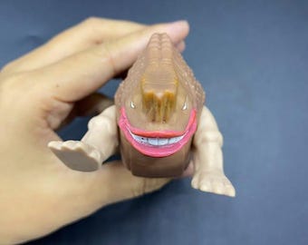Può includere: Un giocattolo mostruoso marrone con una testa strutturata e arti marrone chiaro. Il giocattolo ha un ampio sorriso con labbra rosse e denti bianchi. Il giocattolo è tenuto in una mano su uno sfondo scuro.