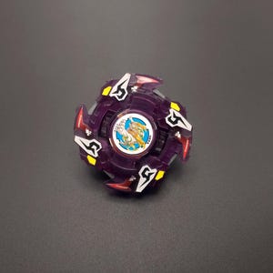 May include: A purple and white Beyblade with a white and yellow design on the centre. The Beyblade has a five-pointed star design on the outer rim.