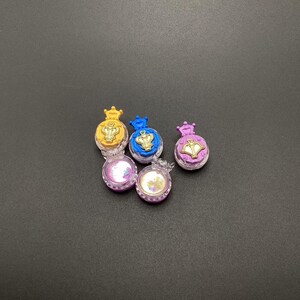 May include: Five small, clear plastic containers with gold crowns and a gold design on the lid. The containers are filled with a glittery substance and are in the colors yellow, blue, purple, and two shades of pink.