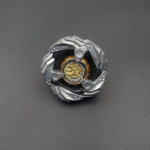 Pu&ograve; includere: Un giocattolo a trottola argentato e nero con un centro dorato raffigurante una grafica di leone e la parola "BEYBLADE". Il giocattolo ha un design multistrato con forma circolare.