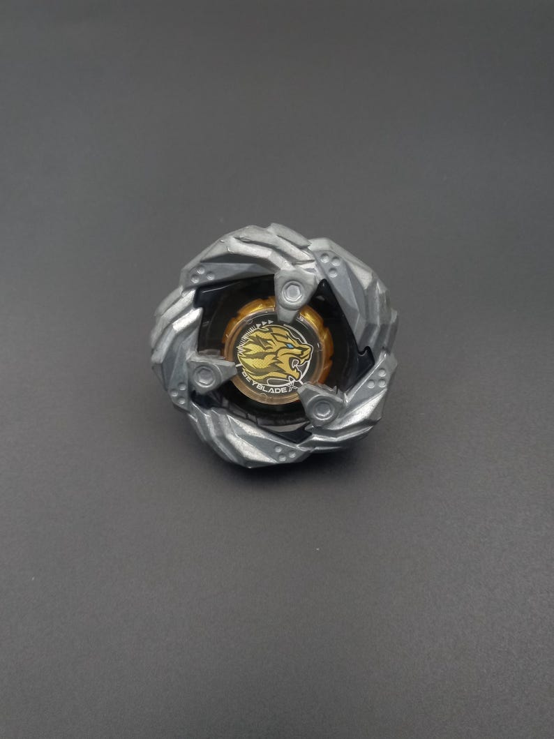Pu&ograve; includere: Un giocattolo a trottola argentato e nero con un centro dorato raffigurante una grafica di leone e la parola "BEYBLADE". Il giocattolo ha un design multistrato con forma circolare.