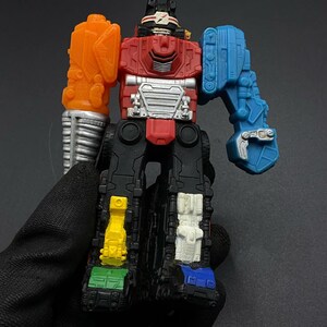 Pode incluir: Um robô de brinquedo colorido com um torso vermelho, braços laranja e azuis e pernas pretas. O robô tem um braço prateado e vários detalhes coloridos, incluindo detalhes amarelos, verdes, brancos e azuis. O brinquedo é segurado por uma mão enluvada.