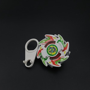May include: A white, red, green, and yellow spinning top toy with a white handle. The toy has a circular design with a central image of a dragon. The handle is white and has a loop and a hook.