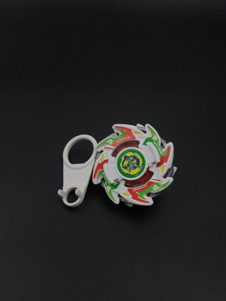 May include: A white, red, green, and yellow spinning top toy with a white handle. The toy has a circular design with a central image of a dragon. The handle is white and has a loop and a hook.