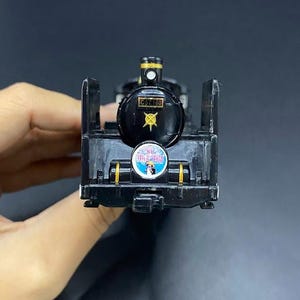 May include: A black toy train with gold and white detailing. The front of the train has the text "C57 180" and a circular emblem. The toy is a miniature replica of a steam locomotive.