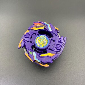 Puede incluir: Una peonza morada con detalles en naranja y azul claro, un emblema circular central con borde amarillo, y el texto "GIA DRAGOON" junto a caracteres japoneses impresos en la superficie superior de la estructura de plástico.