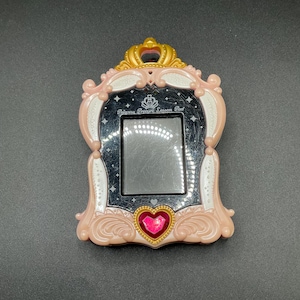 May include: A pink and gold Princess Cure Lesson Pad toy with a crown design. The toy has a rectangular screen, a heart-shaped gem, and the text "Princess Cure Lesson Pad".