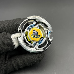 Peut inclure: Un toupie argenté et noir avec un centre jaune et bleu. Le centre présente un graphisme de requin et le mot "Pisces". Le jouet a un design multicouche avec des accents bleus.